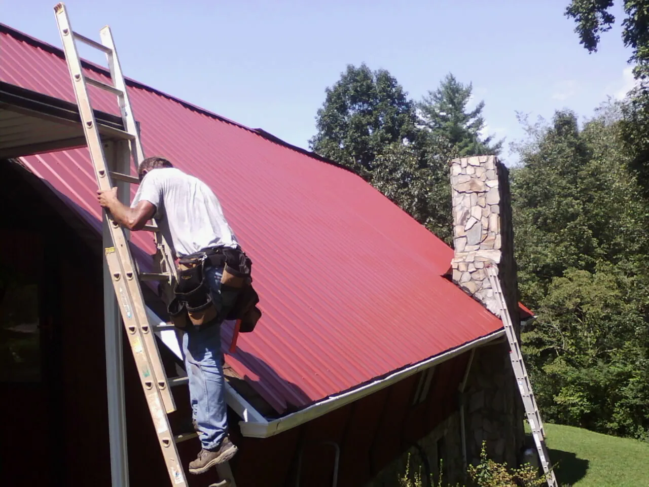 Roofer on a ladder beside a red metal roof and stone chimney — Soffit & Fascia Repair by Roofing Brothers in Black River Falls